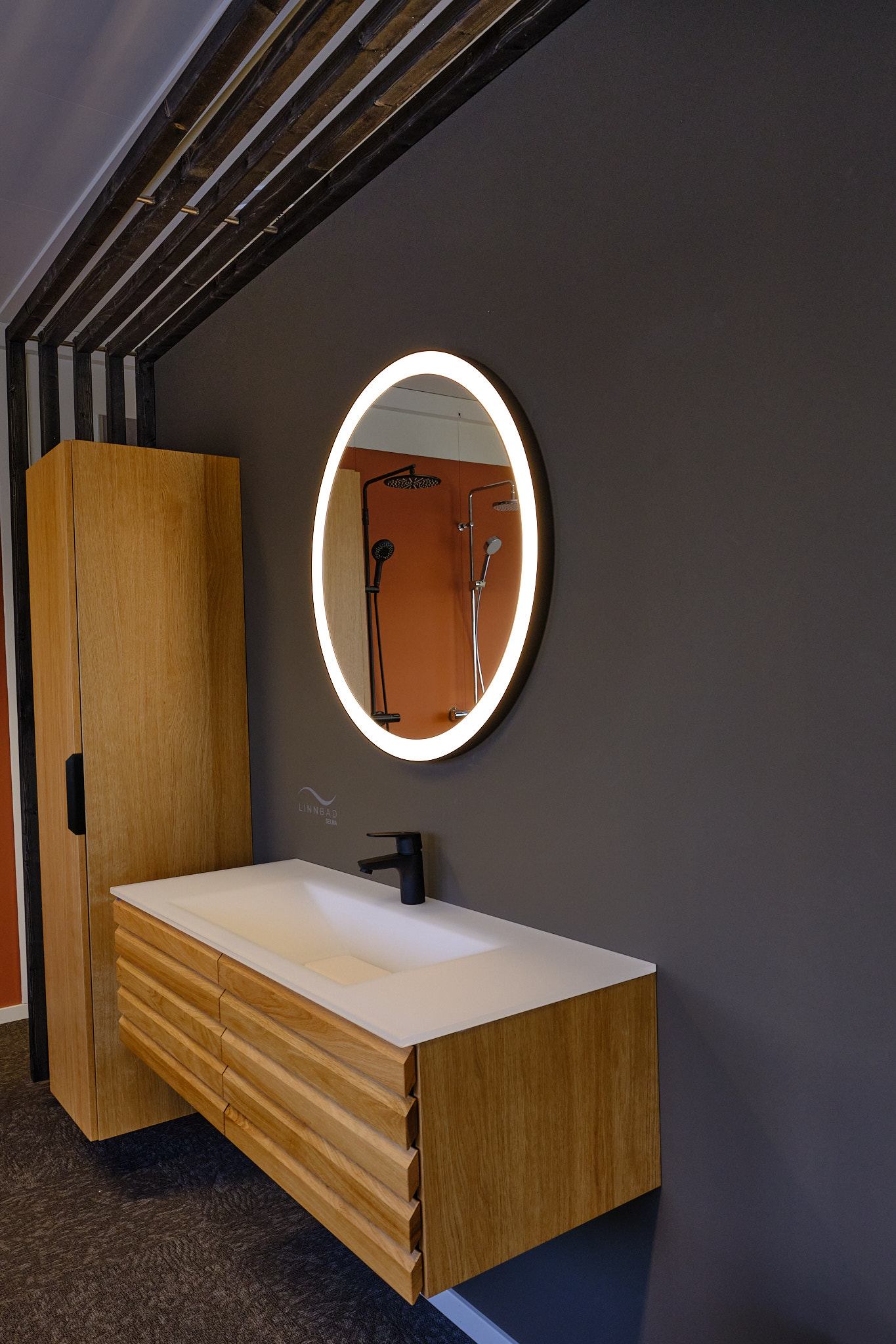 Baderomsmøbel fra Linn Bad i eik med hvit servant og sort servantbatteri. Rundt speil med lys.